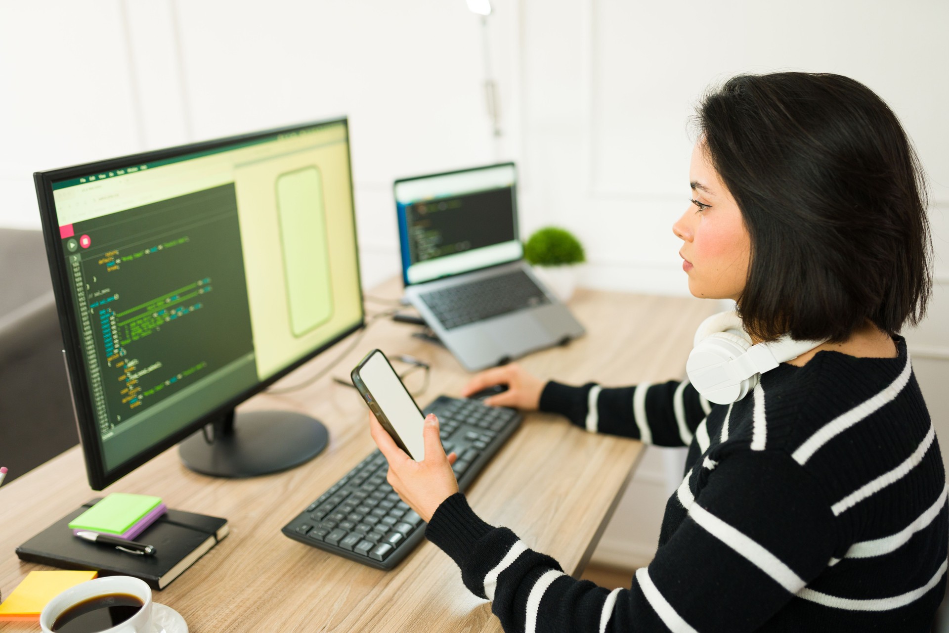 Woman programmer analyzing smartphone screen, comparing mobile data with desktop code during remote work session